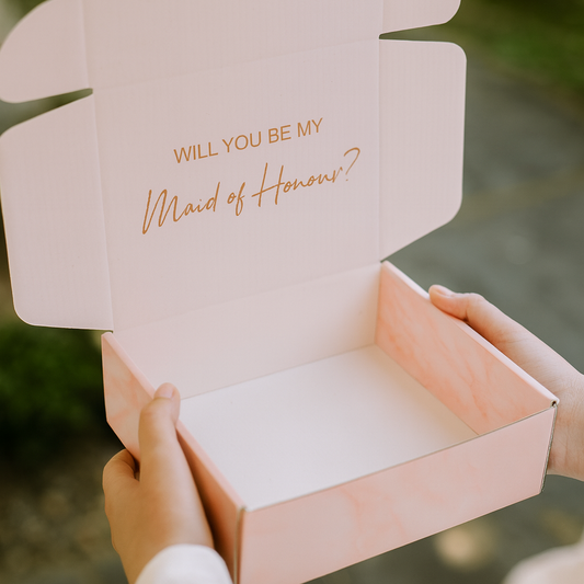 Pink box with 'Will you be my Maid of Honor?' text held by a person.