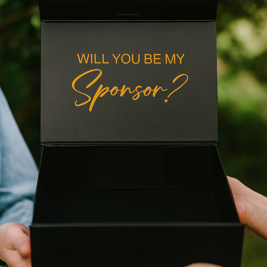 Black box with 'Will you be my sponsor?' text held by two hands against a blurred green background
